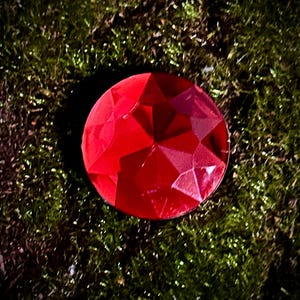 May include: A large, red, faceted gemstone with a shiny, reflective surface. The gemstone is sitting on a bed of green moss.