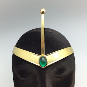 May include: A gold headband with a green gem in the center. The headband has a pointed top and a curved bottom.