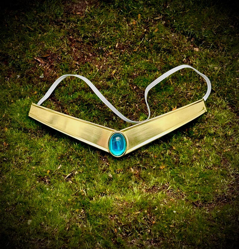 May include: A gold-colored tiara with a blue gem in the center. The tiara is on a green grassy surface.