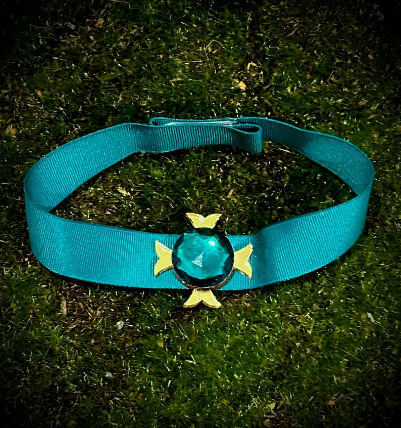 May include: A teal blue ribbon choker with a gold and green jewel pendant. The jewel is a faceted emerald green gemstone.