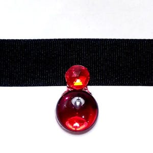 May include: A red crystal choker necklace with a black ribbon. The crystal is faceted and has a silver backing.