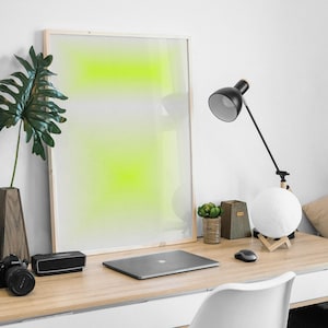 Puede incluir: Un cuadro blanco enmarcado con un fondo degradado verde y blanco se encuentra sobre un escritorio de madera con un portátil, una lámpara y una planta en maceta.