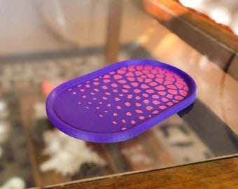 May include: A purple, oval-shaped tray with a raised edge. The tray's surface features a pattern of pink dots and interconnected shapes. The tray is likely made of plastic or a similar material. The design is modern and geometric.