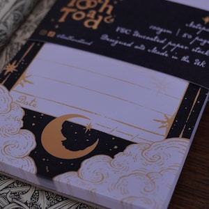 Peut inclure: Un bloc-notes avec un motif noir et or représentant une lune croissante et des nuages. Le bloc-notes contient 50 pages et est fabriqué à partir de papier certifié FSC. Le texte "Tooth & Toa" est imprimé sur la face avant du bloc-notes.