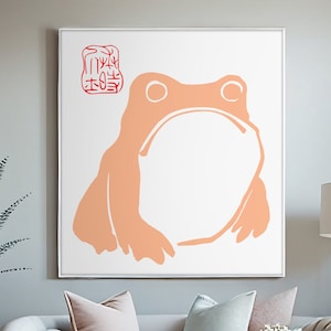 May include: A white framed print of a peach-colored frog with a red stamp in the top left corner. The print is hanging on a gray wall above a white sofa with light blue and peach pillows. A white coffee table with a glass vase and a small plant sits in front of the sofa.