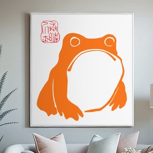 Puede incluir: Una impresión en lienzo blanco con una silueta naranja de una rana. La rana está sentada con las piernas cruzadas y tiene una gran barriga redonda. La impresión está enmarcada en un marco blanco y está colgada en una pared en un salón.