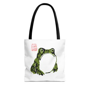 May include: White tote bag with a green and black frog design. The frog has a leaf pattern and a large open mouth. The bag has black handles and a red stamp-like design.