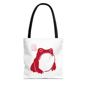 Puede incluir: Bolso tote blanco con una ilustración de rana roja y asas negras. La rana tiene una boca grande y abierta y ojos redondos. Un pequeño diseño rojo tipo sello está en la esquina superior izquierda.