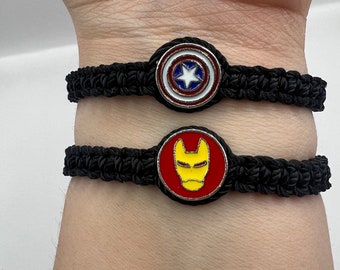 Puede incluir: Dos pulseras trenzadas negras con dijes de metal. La pulsera superior tiene un dije de estrella rojo, blanco y azul. La pulsera inferior tiene un dije de Iron Man rojo y amarillo.