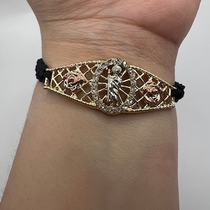 Könnte beinhalten: Ein goldenes Armband mit einem schwarzen Band und einem religiösen Anhänger mit einer Heiligenfigur, die von klaren Strasssteinen umgeben ist. Der Anhänger wird von zwei rosafarbenen Blumen flankiert.