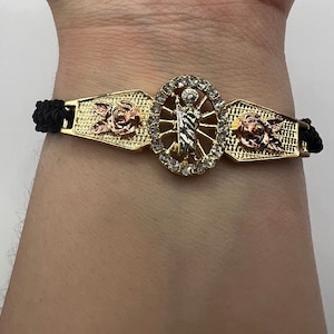 May include: A gold bracelet with a black braided cord. The center of the bracelet features a religious figure with a halo and a rose on each side.