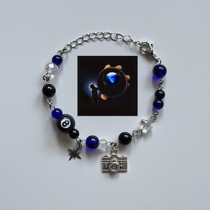 May include: A silver chain bracelet with black and blue beads, clear crystal beads, and a small silver camera charm. The bracelet also features a black bead with an eight ball design and a star charm. An image of a person holding a glowing orb is also included.