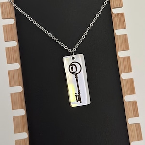 May include: A silver necklace with a rectangular pendant featuring a key design with a letter D in the keyhole. The pendant is on a silver chain.