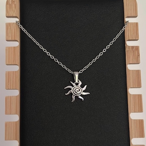 Puede incluir: Collar plateado con un colgante de sol. El colgante tiene un centro en espiral con rayos radiantes. El collar se exhibe sobre una superficie negra, con un soporte de madera en el fondo.