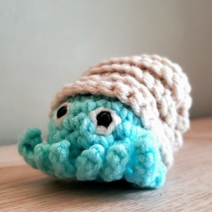 Puede incluir: Un juguete de cangrejo ermitaño de crochet azul y beige con ojos negros.