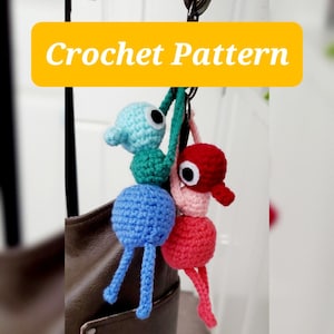 Op de afbeelding: Twee gehaakte figuren hangen aan een metalen ring, de ene lichtblauw en groenblauw, de andere roze en rood, elk met een groot wit oog en zwarte pupil. Een gele banner bovenaan toont de tekst Crochet Pattern in witte letters.