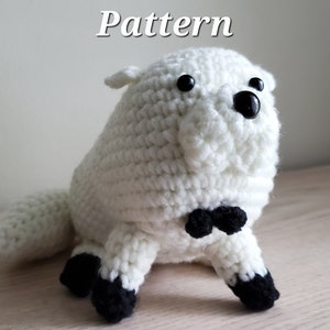 May include: A handmade, white crochet animal with black eyes, a black bow tie, and black feet. The word "Pattern" is written in white at the top of the image. The plush toy is sitting on a wooden surface.