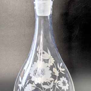 Peut inclure: Carafe en verre transparent avec un bouchon rond et un motif floral gravé. La carafe a un corps bulbeux et un long col. Le motif floral comprend des fleurs et des feuilles. La carafe est probablement utilisée pour servir du vin ou d'autres boissons.