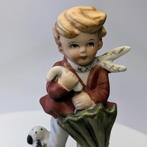 May include: A porcelain figurine of a boy holding an umbrella and a dog. The boy is wearing a red jacket, white pants, and brown shoes. The dog is white with black spots. The figurine is standing on a green base.