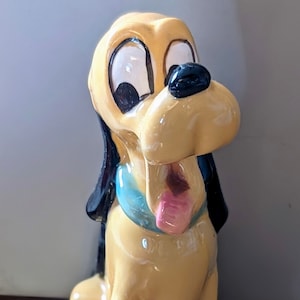 May include: A ceramic figurine of Pluto, a cartoon dog from Disney. Pluto is sitting with his tongue sticking out and has a blue collar. The figurine is yellow with black ears and a black nose.