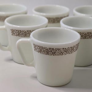 May include: A collection of six white ceramic mugs, each featuring a brown floral border. The mugs have handles and a simple design. They are displayed on a white surface, showcasing their classic style.