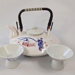 May include: A white ceramic teapot with a dark brown woven handle and a matching lid, featuring a blue and red graphic of a person in a kimono, a bridge, and a pagoda. Two small white ceramic cups with flared rims accompany the teapot.