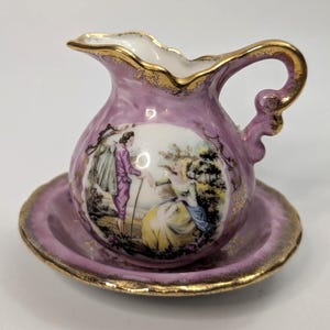 Peut inclure: Un petit pot à lait en porcelaine lavande avec sa soucoupe assortie, orné de bords dorés et d'une scène peinte à la main représentant deux personnages dans un paysage. Le pot présente un bord festonné et une anse courbée avec des détails dorés.