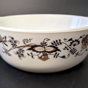 May include: White ceramic bowl with a decorative floral pattern in brown and gold. The bowl has two handles and a rounded shape, suitable for serving. The floral design includes stylized leaves and flowers.