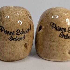 May include: A pair of ceramic salt and pepper shakers shaped like potatoes with a textured tan and brown speckled finish. Each shaker features the black printed text Prince Edward Island on the front and has small indentations resembling potato eyes.