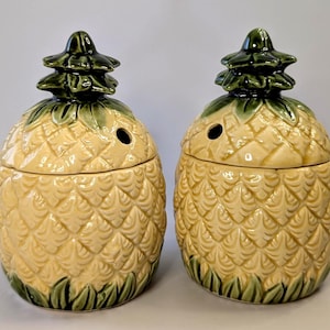 May include: Two ceramic pineapple-shaped canisters with removable lids. Each canister features a cream-coloured textured body with a diamond pattern, a dark green leafy base, and a dark green leafy top handle. Each lid has a small circular hole.