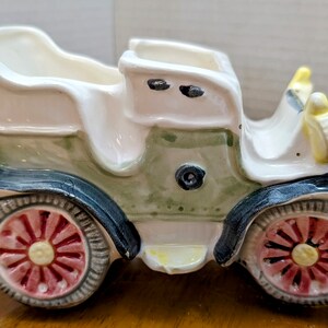 Peut inclure: Un pot de fleurs en céramique vintage de couleur blanche avec des accents verts et noirs. La voiture a des roues rouges avec des centres jaunes.