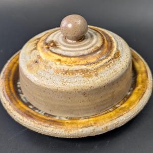Puede incluir: Una mantequera de cerámica con tapa. El plato y la tapa son una mezcla de colores beige, marrón y amarillo-anaranjado. La tapa tiene un pequeño pomo redondo en la parte superior. El plato se asienta sobre un plato a juego.