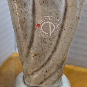 Może przedstawiać: Nakrapiany, jasnoszary wazon z kwadratową podstawą i żłobkowanym, teksturowanym korpusem. Wazon ma etykietę z napisem "Canadiana Pottery Canada". Wazon jest przedmiotem dekoracyjnym.