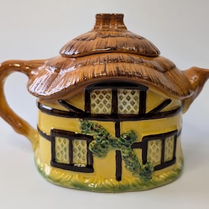 Peut inclure: Une théière en céramique en forme de cottage. Elle présente un couvercle en chaume, des murs jaunes avec des bordures noires et un motif de vigne verte. La poignée et le bec sont bruns. Les fenêtres ont un motif en losange.