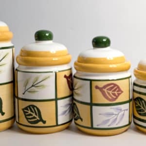 Puede incluir: Juego de cuatro botes de cocina de cerámica con tapas. Los botes son blancos, amarillos y verdes, con diseños de hojas. Cada bote tiene un pomo verde en la tapa. Los botes son de diferentes tamaños.