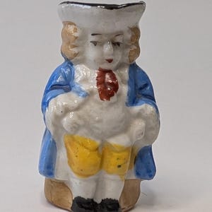 Peut inclure: Petite figurine vintage représentant un homme en costume du XVIIIe siècle. La figurine porte un manteau bleu, une culotte jaune et un chapeau blanc. La figurine a une cravate rouge et des chaussures noires. Le design est détaillé et probablement en céramique.