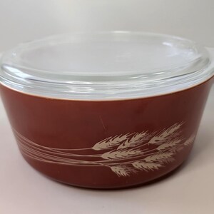 Könnte beinhalten: Eine runde, kastanienbraune Pyrex-Auflaufform mit einem Deckel aus klarem Glas. Das Vintage-Geschirr zeigt ein Weizenmuster in Creme. Die Schüssel hat zwei Griffe und ist für den Ofen geeignet. Ein klassisches Küchenutensil.