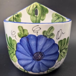 Op de afbeelding: Een witte keramieken wandpot met een grote, handgeschilderde blauwe bloem in het midden, omringd door groene bladeren en zwarte krullende lijnen. Het object heeft een driehoekige achterplaat met een ophanggat en een blauwe rand.