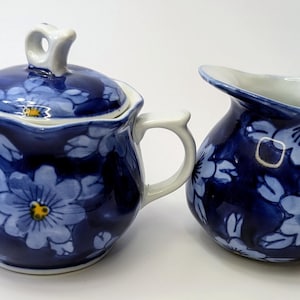 Peut inclure: Un ensemble sucrier et crémier en porcelaine bleu et blanc avec un motif floral. L'ensemble comprend un sucrier avec couvercle et une anse, ainsi qu'un pot à crème avec une anse.