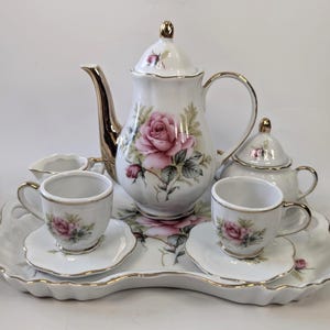 May include: A white porcelain tea set with a floral rose design. The set includes a teapot, sugar bowl, creamer, two teacups with saucers, and a serving tray. Gold trim accents the edges of each piece.