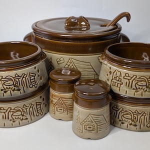 May include: A brown and beige ceramic soup tureen set with a lid, ladle, and several bowls. The set includes two salt and pepper shakers. The bowls and shakers have a decorative design of a house and other elements.