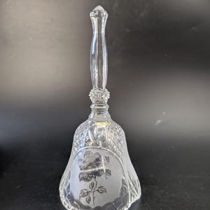 May include: A clear glass bell featuring a tall, slender handle with a textured, faceted base. The bell body displays a frosted finish with an etched rose design on the front, surrounded by a diamond-cut pattern and a scalloped bottom edge.