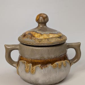 May include: A ceramic sugar bowl with two side handles and a matching lid. The vessel features a two-tone glaze with a smooth grey upper section and a textured, drippy white and brown lower section. The lid has a rounded knob handle.
