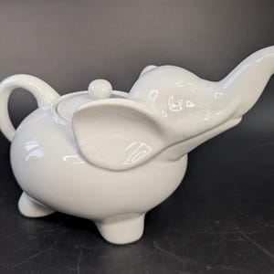 Peut inclure: Théière blanche en céramique en forme d'éléphant. Elle a un bec verseur incurvé, une anse ronde et un petit couvercle avec un bouton. La trompe de l'éléphant forme le bec, et ses oreilles sont visibles sur les côtés.