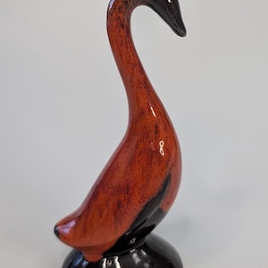 Może przedstawiać: Ceramiczna figurka łabędzia z długą, zakrzywioną szyją i błyszczącym wykończeniem. Korpus łabędzia jest czerwonobrązowy z czarnymi akcentami i stoi na czarnej podstawie. Figurka jest przedmiotem dekoracyjnym.