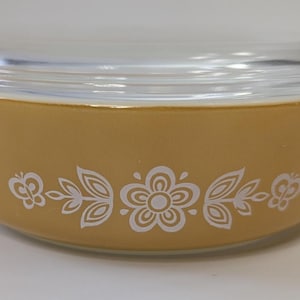 Könnte beinhalten: Eine vintage, senffarbene Pyrex-Schüssel mit einem Deckel aus klarem Glas. Die Schüssel hat ein weißes Blumenmuster auf der Außenseite. Die Schüssel hat zwei Griffe und ist für den Ofen geeignet. Ein klassischer Küchenartikel.