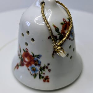May include: A white ceramic bell with a floral pattern and a gold cord. The bell has small holes around the bottom.