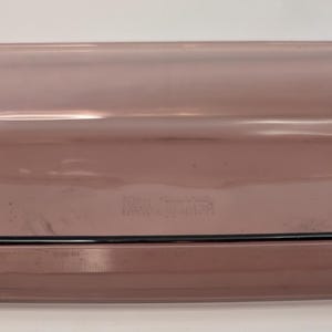 Puede incluir: Una fuente rectangular de vidrio transparente, con esquinas redondeadas y un borde negro. La fuente tiene dos asas y es de color morado claro. Este utensilio de cocina es adecuado para hornear guisos, postres y otros platos.