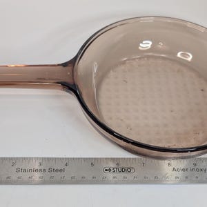 May include: A round, transparent, light brown glass frying pan with a single handle. A stainless steel ruler is positioned along the bottom edge, showing the pan has a diameter of approximately 20 cm and a total length of 28 cm.