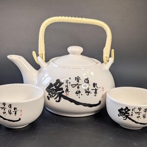 May include: White ceramic tea set with black calligraphy. The set includes a teapot with a woven handle and two matching teacups. The teapot and cups feature decorative black calligraphy.